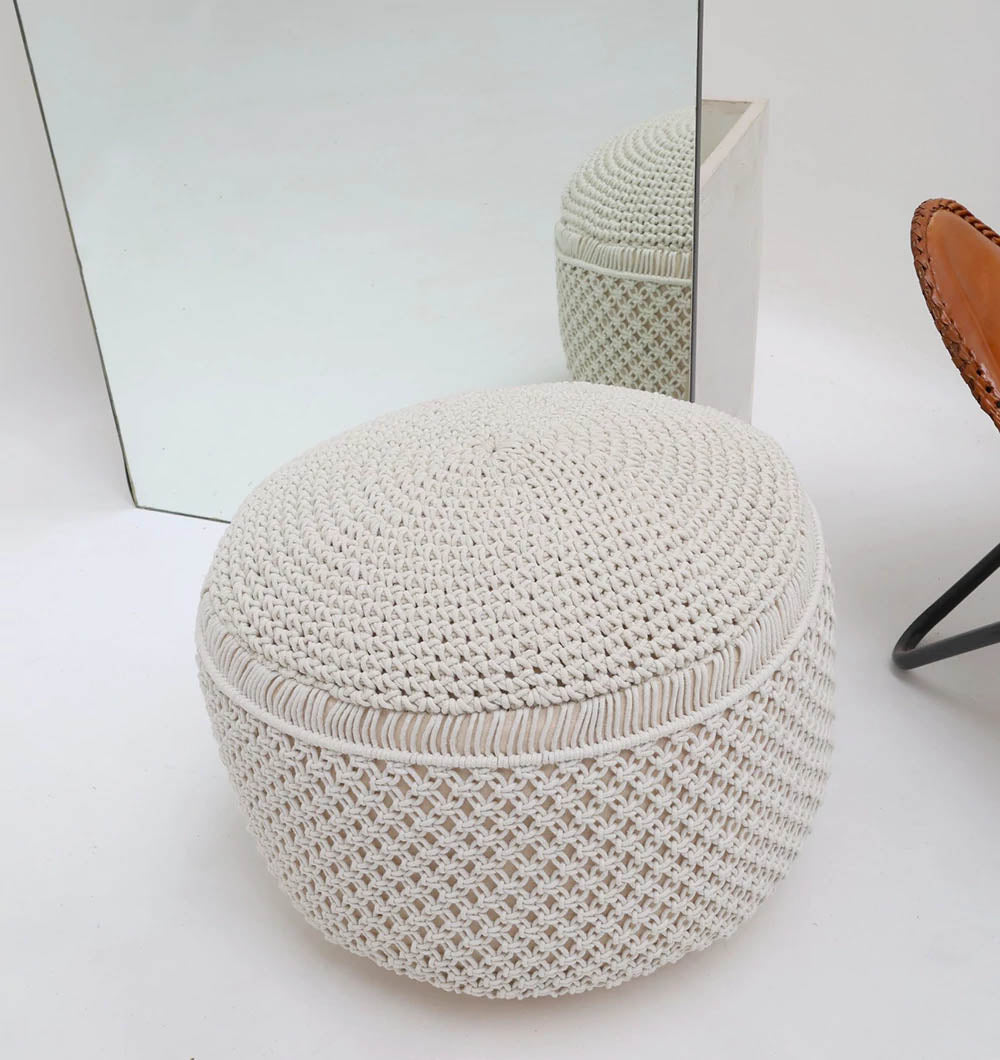 The Boho Macrame Pouf White Round PF-158 by RAMSHA HOME sits on a white floor near a large mirror reflecting the pouf; part of a brown leather chair with black metal legs is visible on the right.
