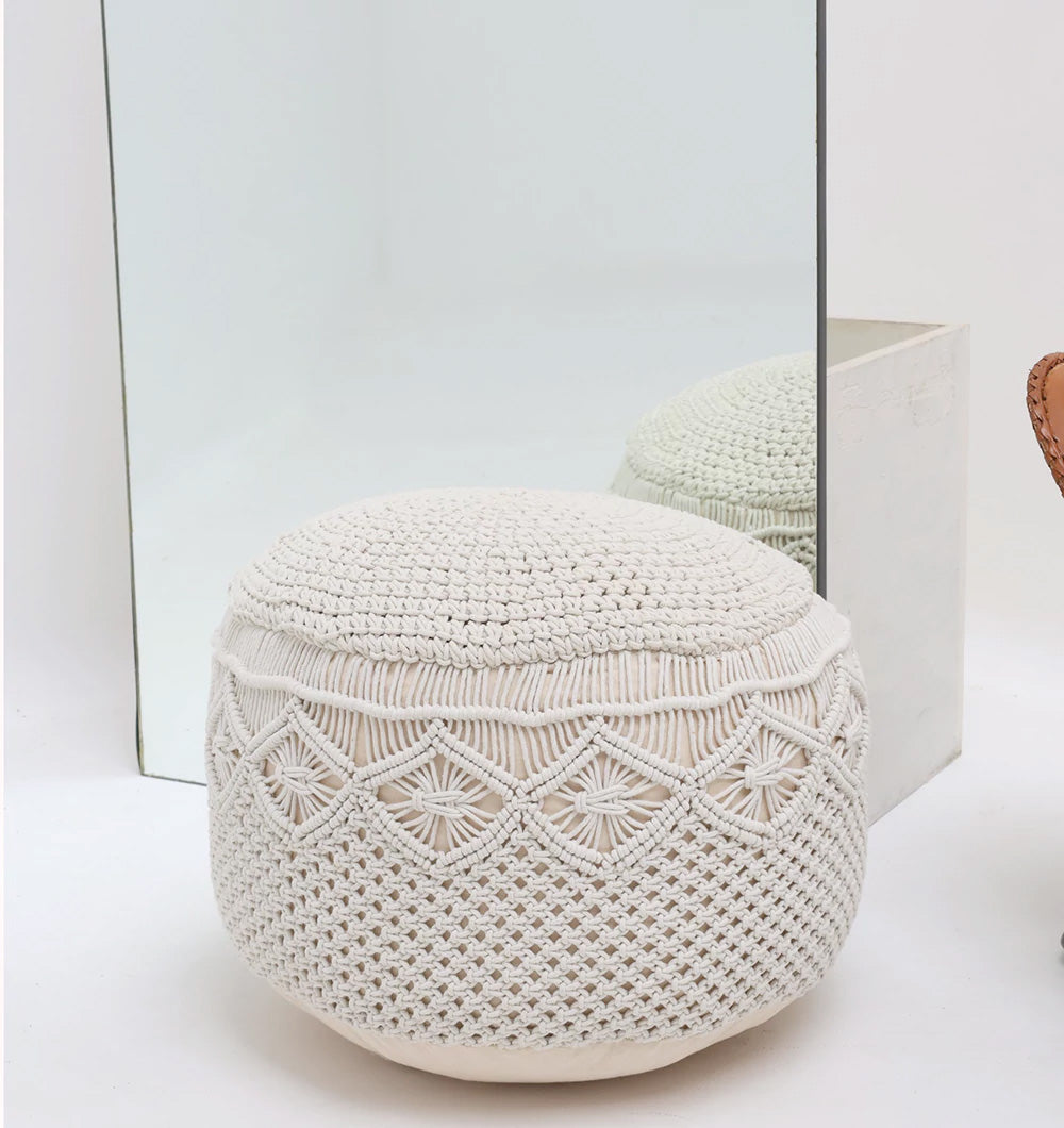 The RAMSHA HOME Boho Macrame Pouf White Round PF-159 sits on a white floor in front of a large mirror, reflecting its textured pattern. A wicker chair is partly visible on the right side of the image.