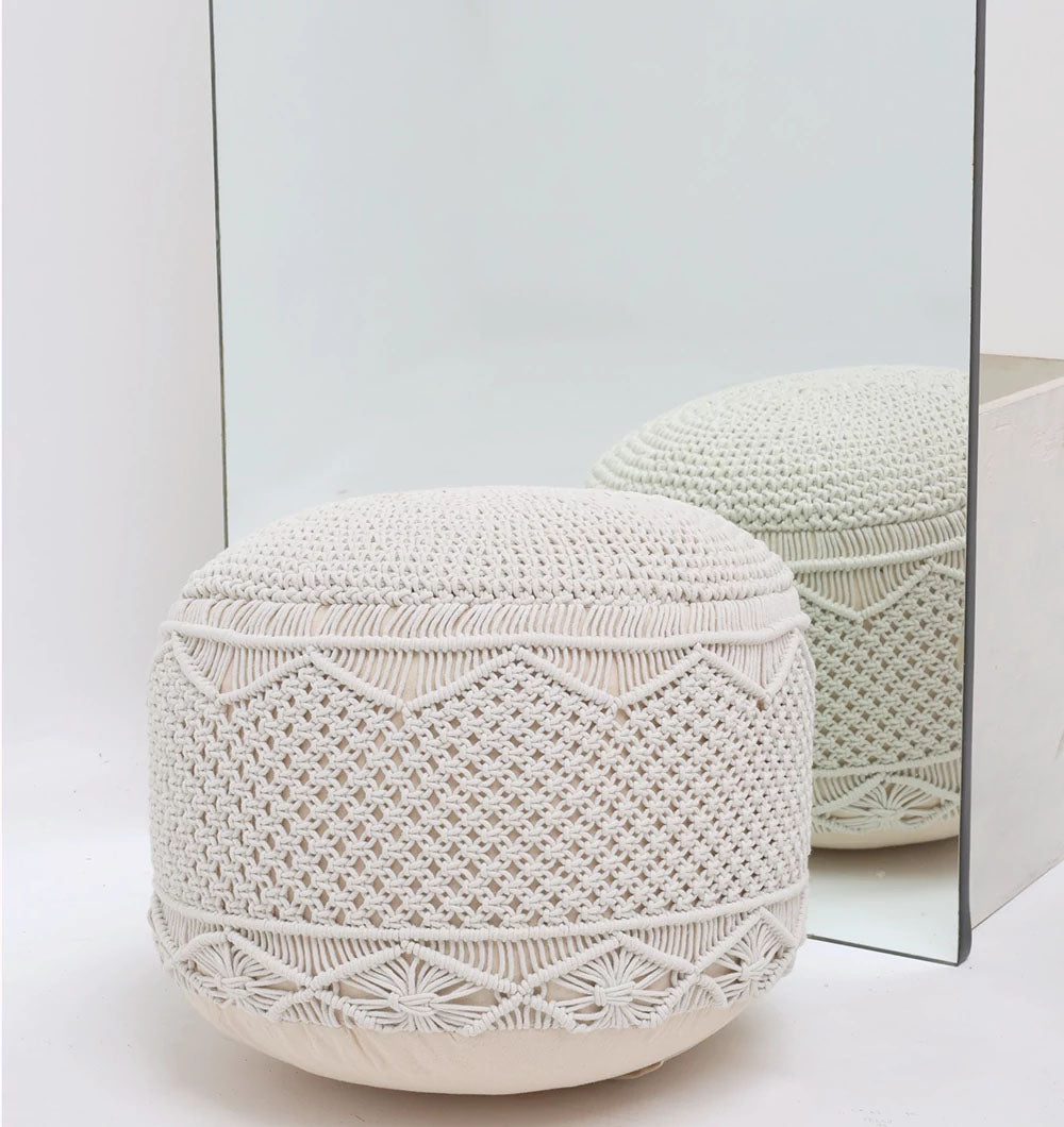 The Handwoven White Macrame Pouf with Bean Bag Insert PF 160 by RAMSHA HOME, featuring intricate cream-colored patterns, sits next to a mirror in a minimal white setting, its textured design beautifully reflected.