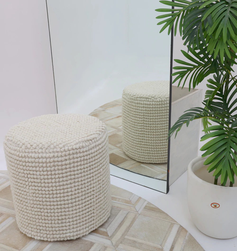 The RAMSHA HOME Handwoven White Macrame Pouf with Bean Bag Insert PF 161 sits on a patterned floor by a large mirror that reflects it. Nearby, a green plant in a white pot adds to the minimal, modern décor.