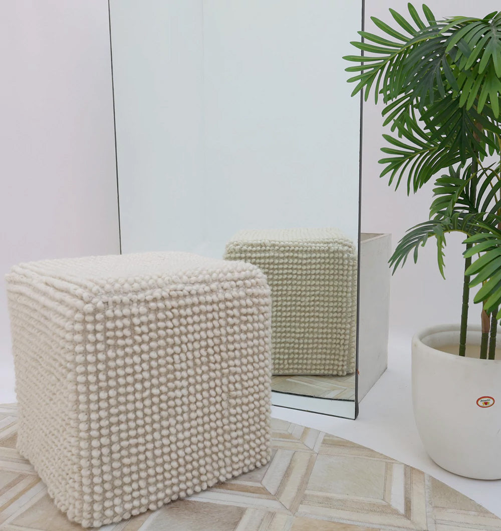 The Handwoven White Macrame Pouf with Bean Bag Insert PF 162 by RAMSHA HOME sits on a tiled floor beside a large mirror and a potted green plant in a white pot, with the pouf’s reflection visible in the mirror.