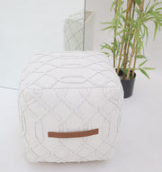 The RAMSHA HOME Ivory Nordic Bliss Cotton Pouf with insert PF 167, featuring a brown leather handle, rests on a white floor near a potted green plant and a mirror that reflects both the pouf and the vibrant foliage.