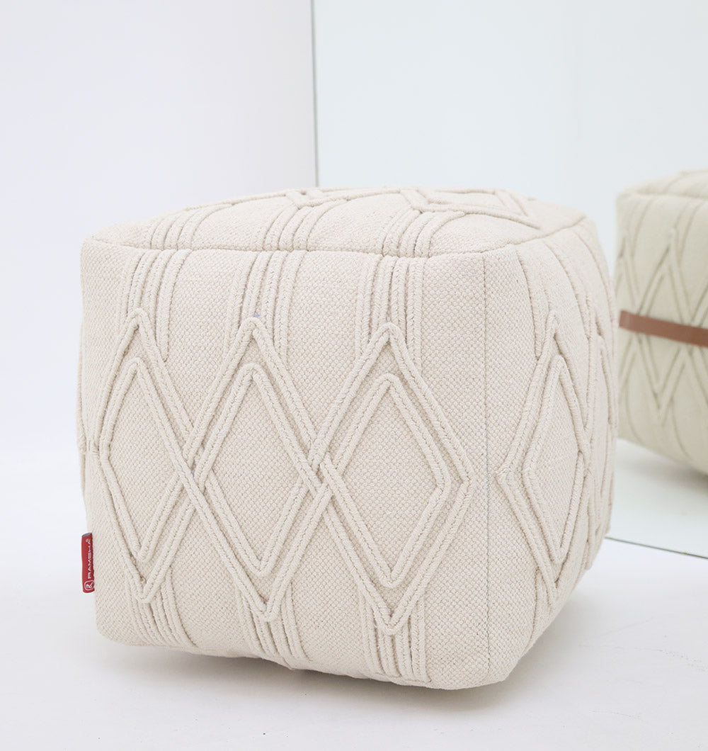 The RAMSHA HOME Ivory Nordic Bliss Cotton Pouf with insert PF 168, cube-shaped with geometric diamond patterns, sits on a white surface. Its textured cotton and soft hues are reflected in a mirror behind it.