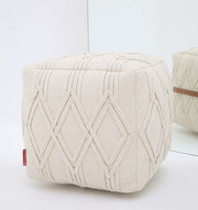 The RAMSHA HOME Ivory Nordic Bliss Cotton Pouf with insert PF 168, cube-shaped with geometric diamond patterns, sits on a white surface. Its textured cotton and soft hues are reflected in a mirror behind it.