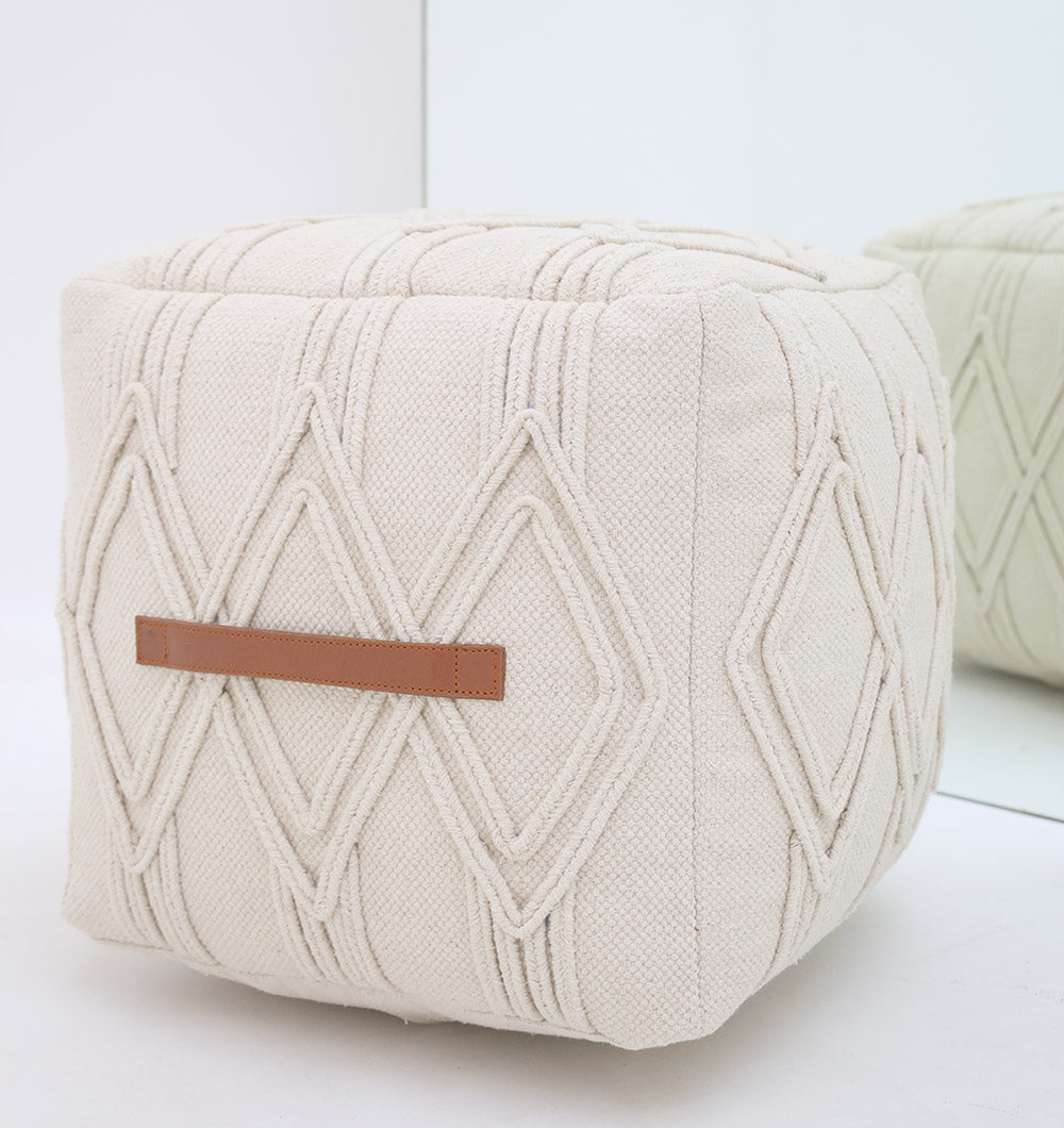 The RAMSHA HOME Ivory Nordic Bliss Cotton Pouf with insert PF 168, featuring a cream hue, textured diamond patterns, and a leather strap, sits on a white floor by a mirror reflecting its side.