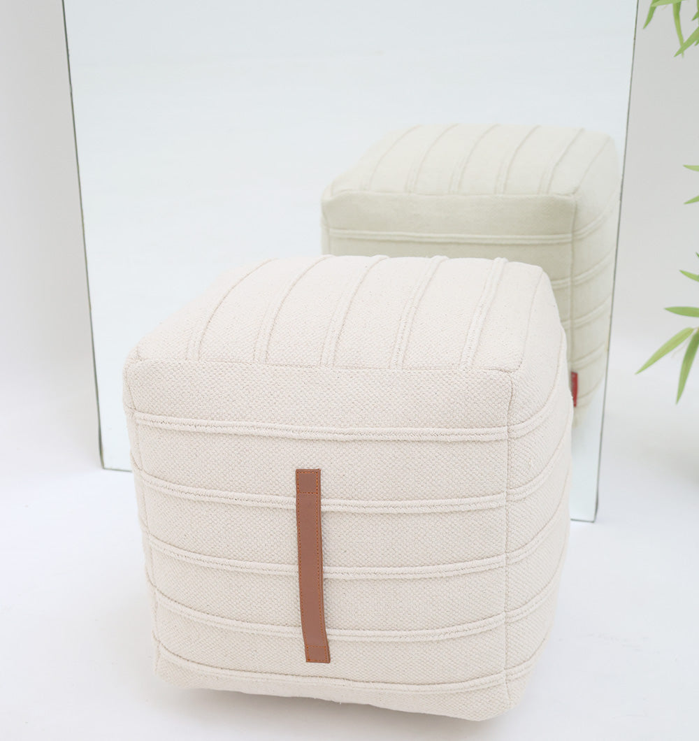 The Ivory Nordic Bliss Cotton Pouf with insert PF 169 by RAMSHA HOME, featuring a textured cream finish and vertical brown strap, sits before a mirror reflecting its stylish look, complemented by green leaves for a fresh decorative accent.