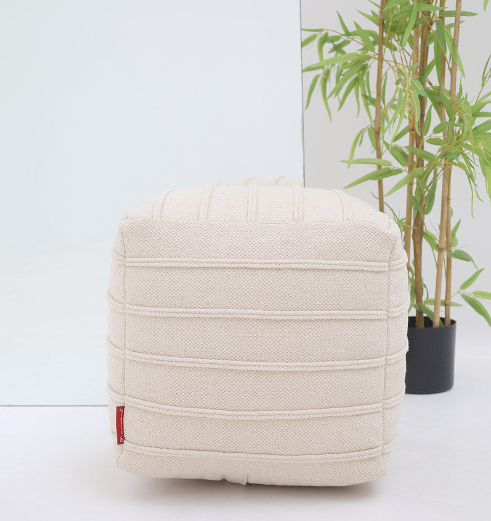 The RAMSHA HOME Ivory Nordic Bliss Cotton Pouf with insert (PF 169) sits before a mirror as a decorative accent. In the background is a potted bamboo, and a small red tag appears on the pouf’s lower left corner.