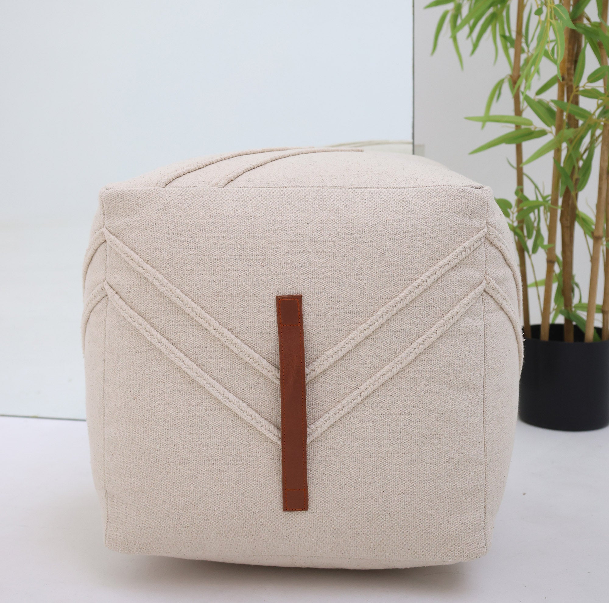 The RAMSHA HOME Ivory Nordic Bliss Cotton Pouf with insert PF 172, featuring a leather handle, adds an elegant accent to any room when placed near a potted plant and mirror on a white floor.