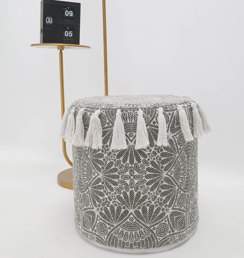 The RAMSHA HOME Stylish cotton printed pouf with decorative tassels PF 174, featuring a black and white geometric design, sits in front of a gold side table with a retro flip clock, all against a plain white background.
