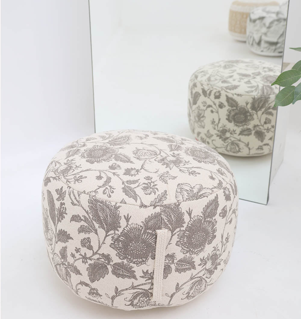 The RAMSHA HOME Round Bohemian Printed Pouf with Comfortable Insert PF 177, featuring a beige and gray floral design, provides decorative seating in front of a mirror. Another pouf and a green plant appear in the background.