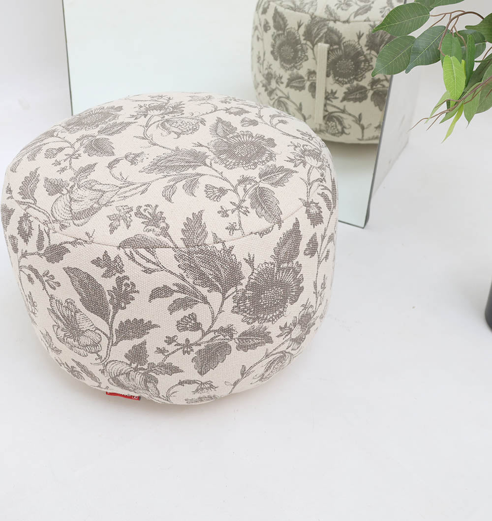 The RAMSHA HOME Round Bohemian Printed Pouf with Comfortable Insert PF 177, featuring a gray pattern, sits on a white floor by a mirror and green plant, reflecting its stylish and decorative seating design.
