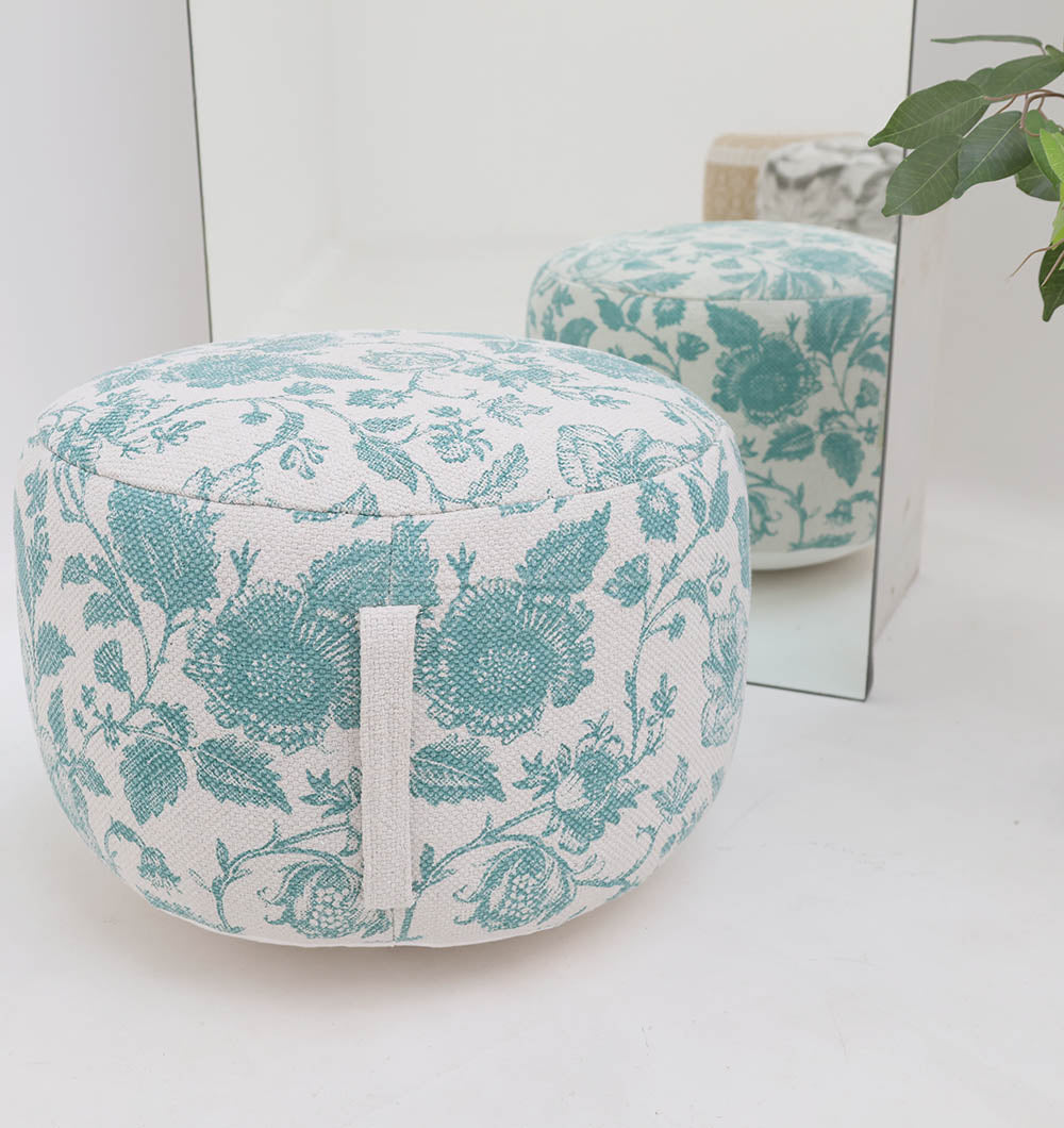 The RAMSHA HOME Round Bohemian Printed Pouf with Comfortable Insert PF 177 features a blue floral design and sits before a mirror, reflecting its vibrant pattern. A green plant in the corner adds freshness to this stylish seating setup.