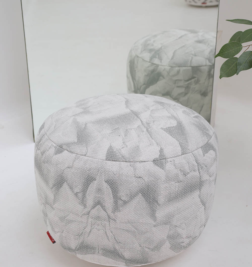 The RAMSHA HOME Round Bohemian Printed Pouf with Comfortable Insert PF 182, in light gray with an abstract pattern, sits on a white floor before a mirror that reflects the pouf and part of a green leafy plant.