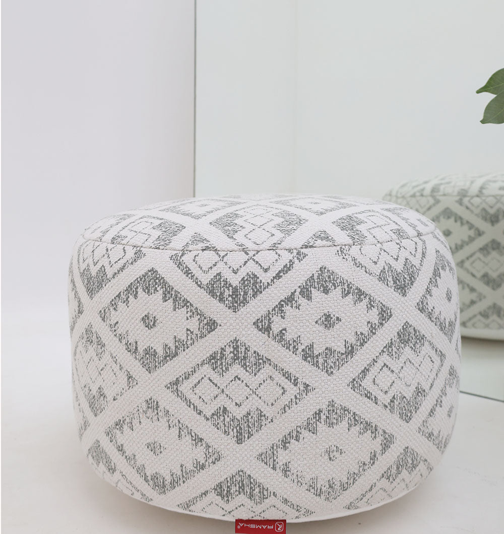 The RAMSHA HOME Round Bohemian Printed Pouf with Comfortable Insert PF 183, featuring a white and gray geometric diamond print, sits beside a mirror on a light floor. A green plant nearby adds freshness, creating a chic, modern décor accent.