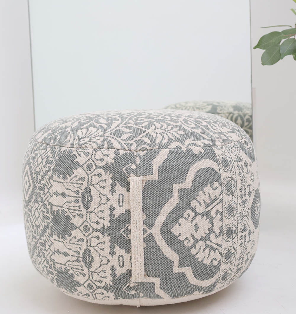 The RAMSHA HOME Bohemian Grey Color Printed Pouf with Comfortable Insert PF 189 sits in front of a mirror, its stylish design reflected. Ideal for home décor, complemented by greenery from a plant on the right side.