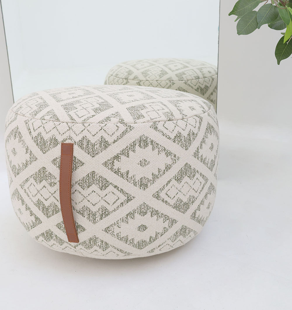 The RAMSHA HOME Luxury Round Floor Pouf with Leather Handle PF 190, featuring a cream geometric diamond print and brown leather handle, rests on a white floor by a mirror with green leaves visible in the upper right corner.