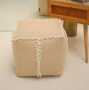 A square jute floor pouf with fringes from RAMSHA HOME (PF 193) sits on a light floor beside a wooden side table with a cane drawer, making for a charming accent piece in the room.