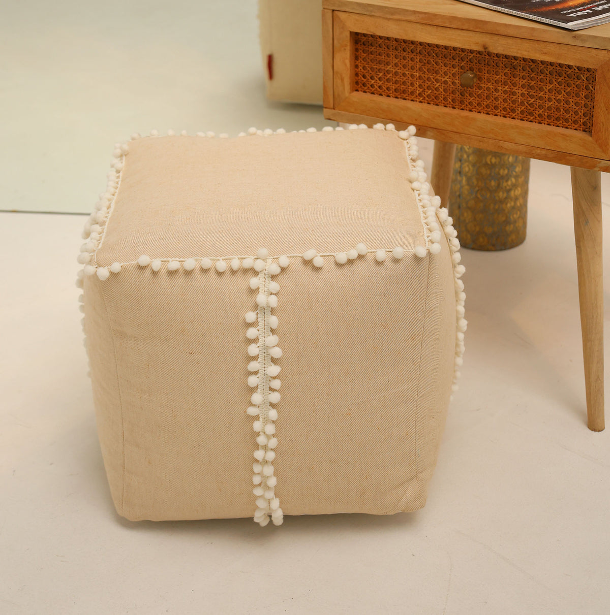 A square jute floor pouf with fringes from RAMSHA HOME (PF 193) sits on a light floor beside a wooden side table with a cane drawer, making for a charming accent piece in the room.