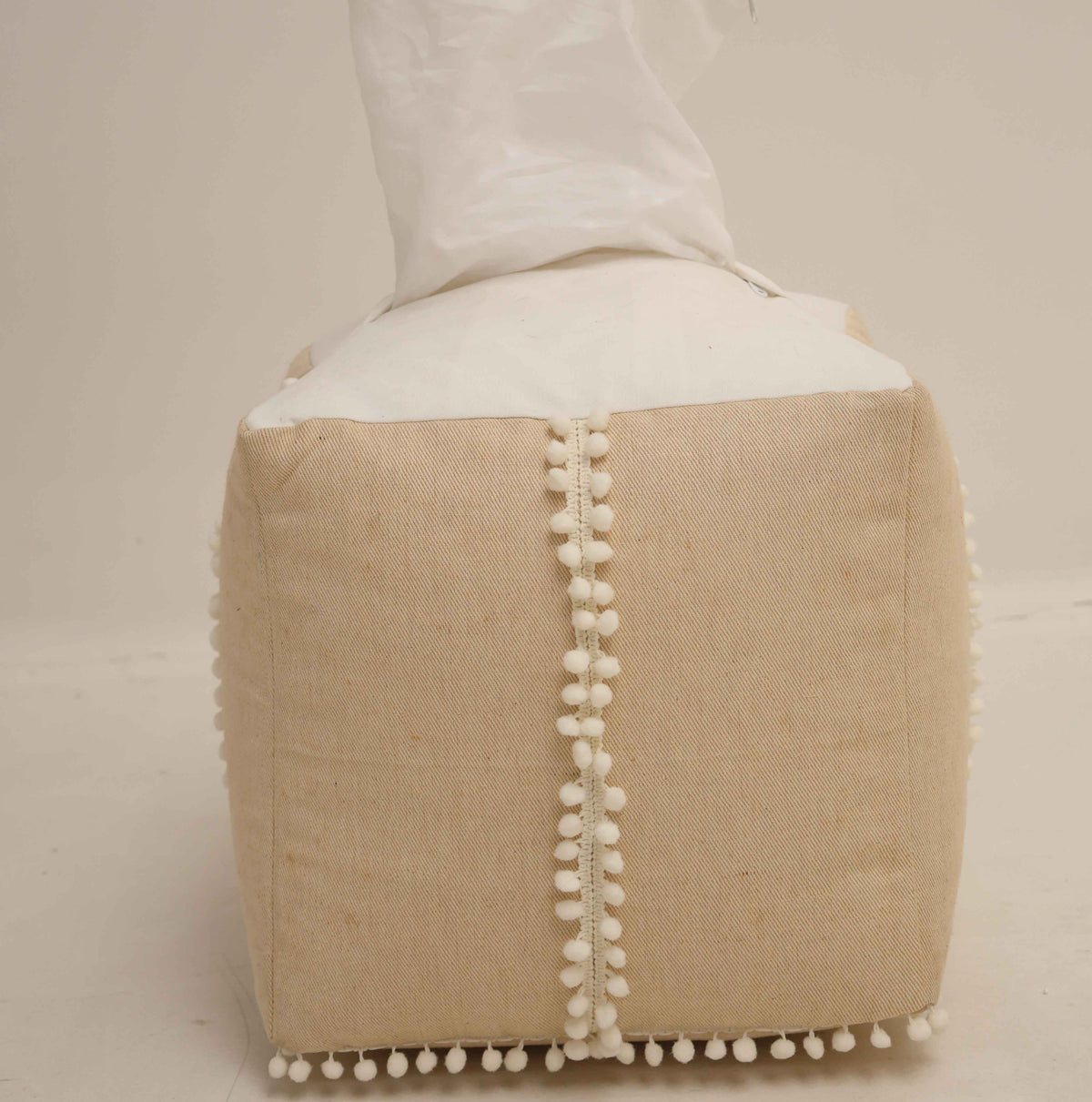 A beige fabric tissue box cover with white pom-pom trim along the seams, holding tissues, placed on a neutral background—perfect as an accent piece to complement your RAMSHA HOME Square Jute Floor Pouf with Fringes PF 193 in any living space.