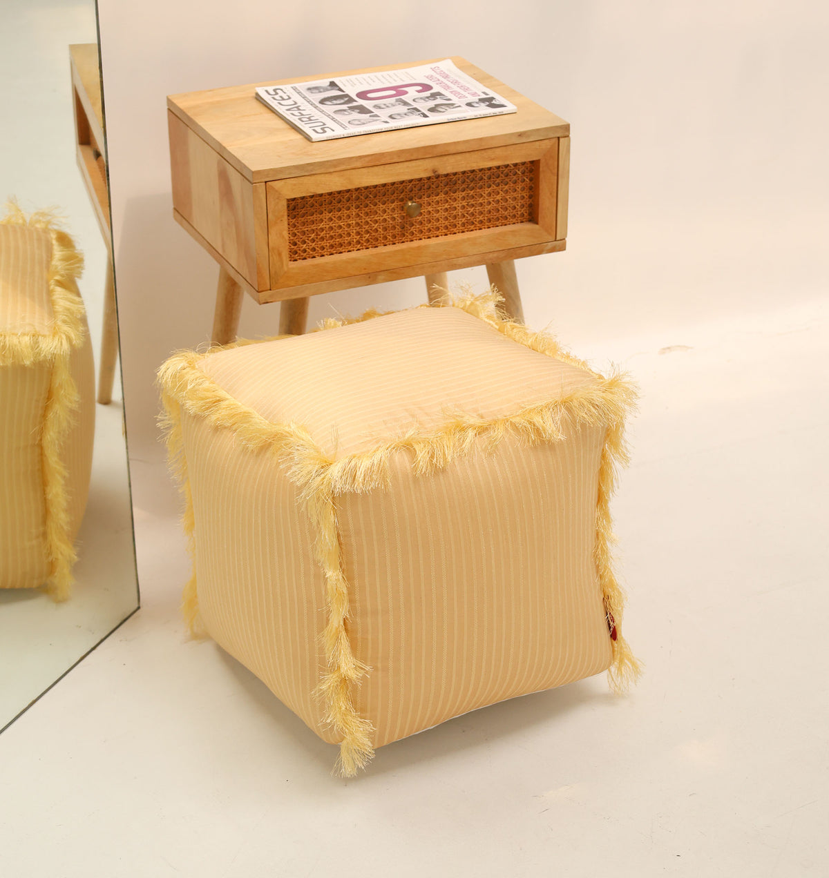 A RAMSHA HOME Square Floor Pouf with Fringes PF 191 in yellow, featuring a cube shape and modern design, is placed beside a wooden side table topped with a magazine and drawer, with the scene reflected in a mirror on the left.