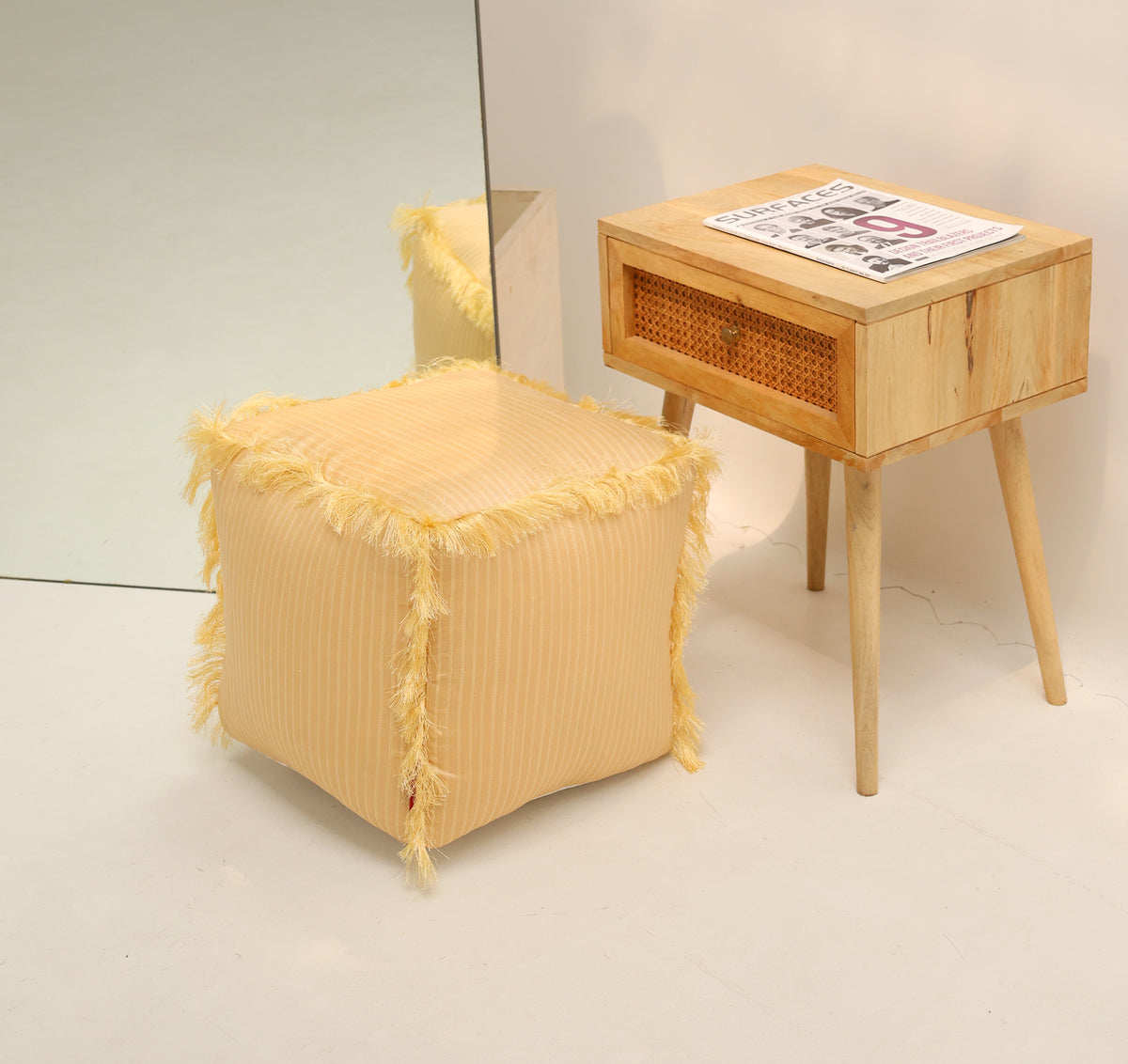 A yellow square floor pouf with fringes by RAMSHA HOME (Square Floor Pouf with Fringes PF 191) sits beside a wooden nightstand with a rattan drawer holding a newspaper, while a mirror behind the pouf reflects both pieces.