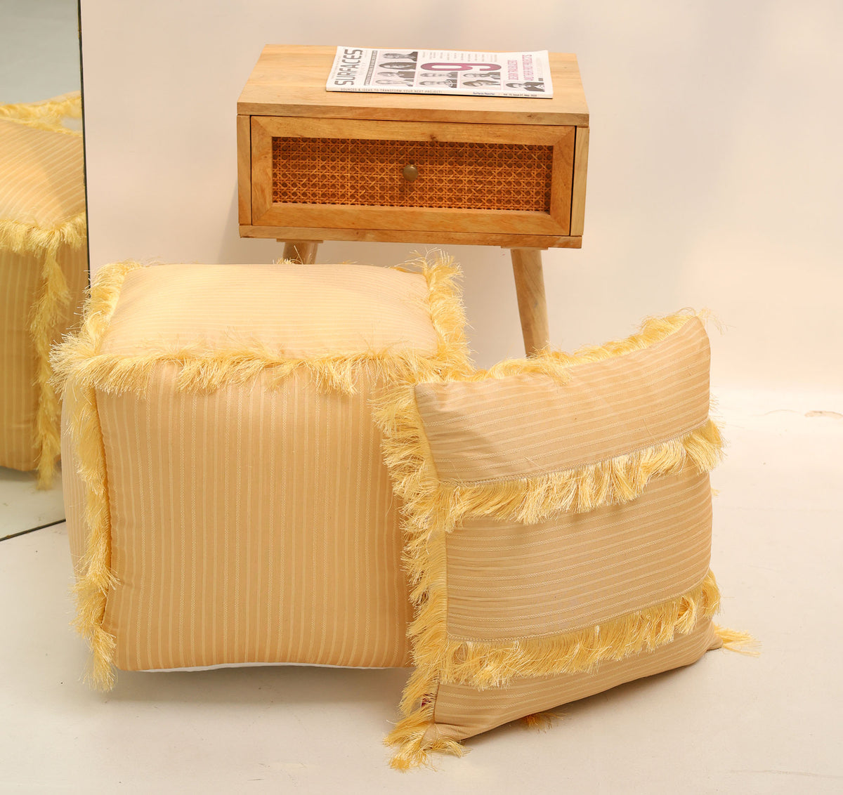 A wooden side table with a drawer holds a newspaper. In front, the RAMSHA HOME Square Floor Pouf with Fringes PF 191, featuring yellow stripes and matching fringe, pairs with a coordinating pillow to bring color and texture to the space. A modern pouf adds contemporary flair to the setting, while a mirror reflects part of the scene.