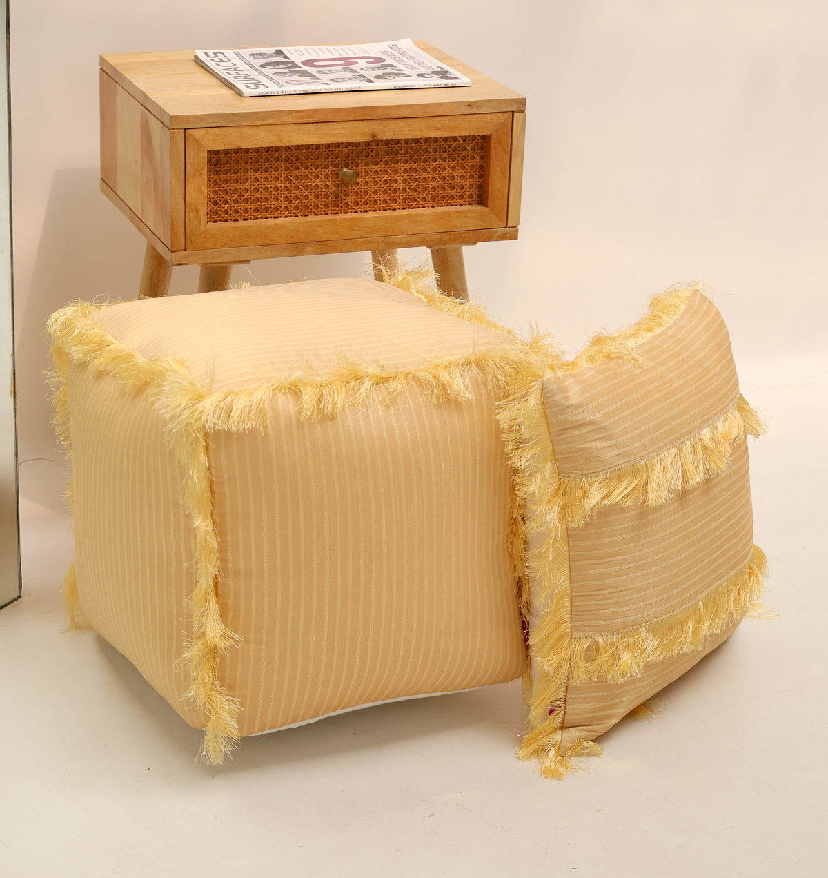 Two yellow, striped Square Floor Poufs with Fringes PF 191 from RAMSHA HOME are placed on the floor beside a small wooden table with a woven drawer and a newspaper on top, set against a plain white background.