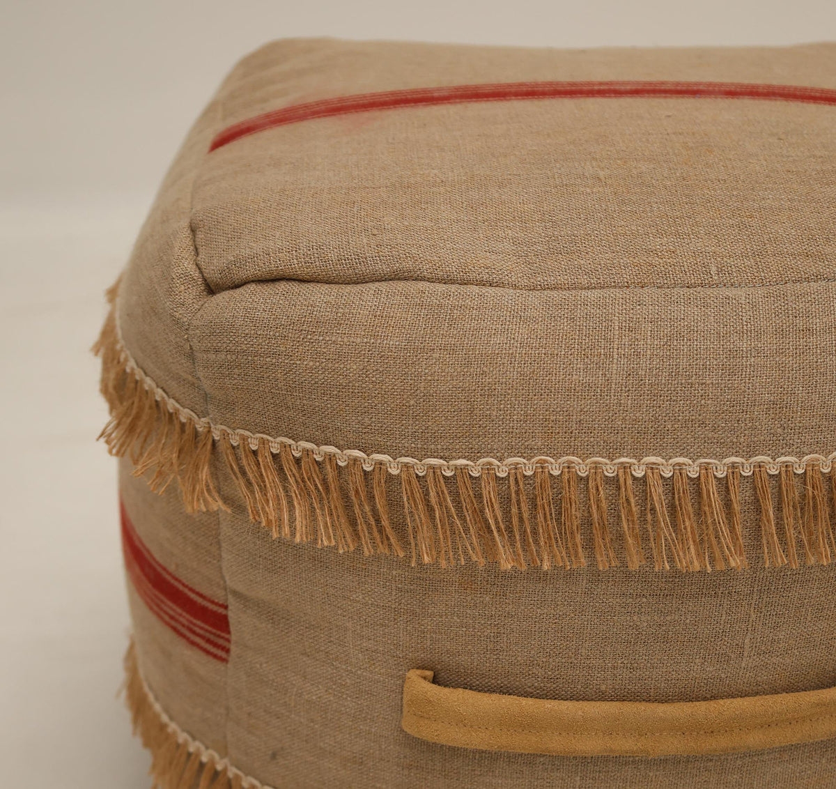 A close-up of the RAMSHA HOME Square Jute Floor Pouf with Fringes PF 192 shows its beige jute fabric accented by bold red stripes, tan fringe trim, and a rectangular tan side handle. This charming accent piece infuses warmth and artisanal character into your space, with its neutral tones and handcrafted look reminiscent of a classic jute pouf.
