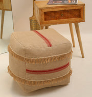 The Square Jute Floor Pouf with Fringes PF 192 by RAMSHA HOME, featuring a beige jute construction with red stripes and decorative fringe trim, is placed in front of a wooden nightstand with a drawer, beside a mirror and decorative broom on a light-colored floor, serving as a charming accent piece.