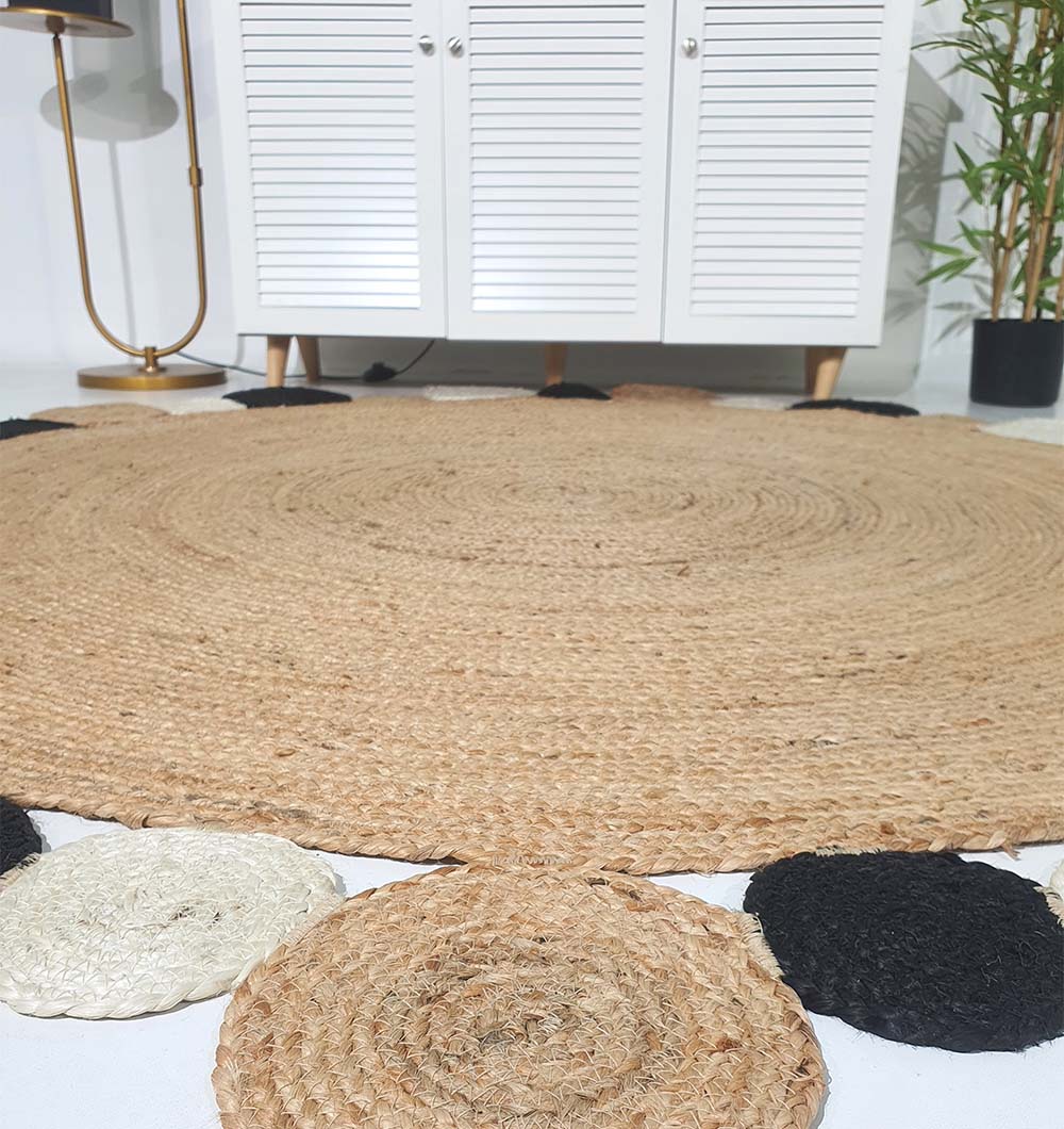 The RAMSHA HOME Jute Natural Rug Round BR-029, featuring braided jute and black and white circular edging, brings boho charm to your space when placed on a light floor near a white cabinet, gold lamp, and potted plant.
