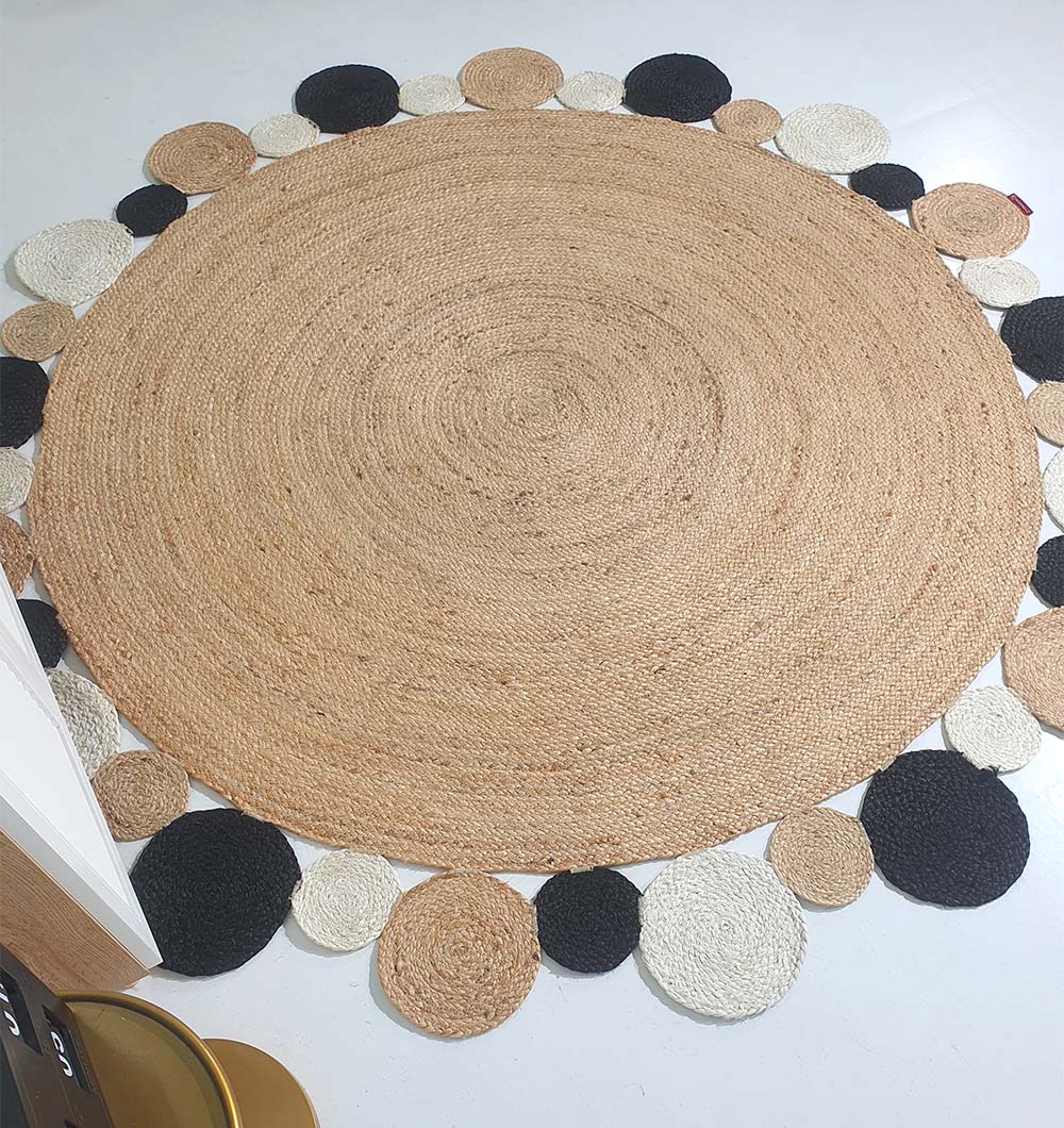 The RAMSHA HOME Jute Natural Rug Round BR-029 features a tan center and alternating black, white, and tan borders for a boho vibe. This braided jute rug adds texture and style, perfect for brightening up any white floor.