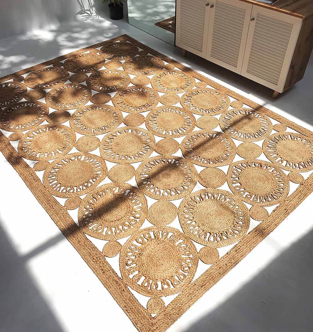 The RAMSHA HOME Braided Natural Outdoor Jute Rug (BR 086), featuring circular patterns and intricate openwork, adds eco-friendly luxury to your white floor near a light cabinet, sunlight highlighting its durable, elegant design.