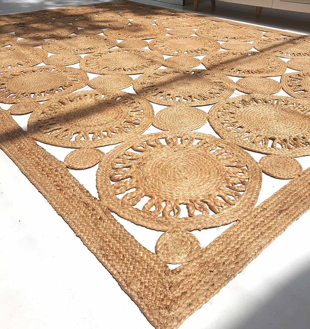 The Braided Natural Outdoor Jute Rug by RAMSHA HOME (BR 086), featuring luxurious, eco-friendly durability and elegant circular and oval patterns, rests on a white floor with sunlight highlighting its textured open weave.