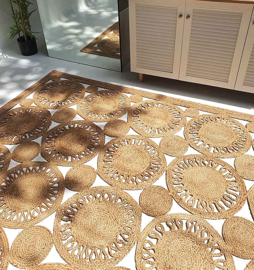 The RAMSHA HOME Braided Natural Outdoor Jute Rug (BR 086), with circular patterns, sits on a white floor by a cabinet and mirror, its woven surface highlighted by dappled sunlight and plant shadows in the bright room.