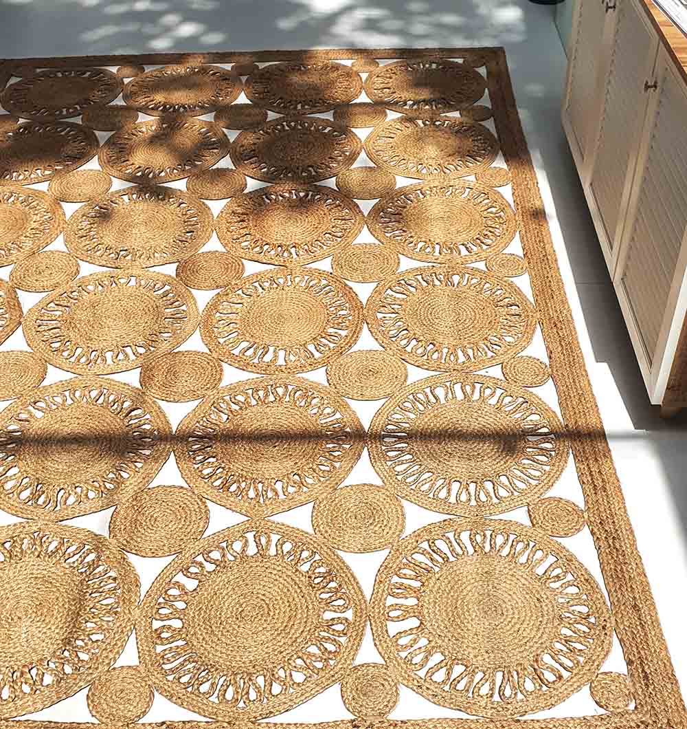 The RAMSHA HOME Braided Natural Outdoor Jute Rug (BR 086) showcases a geometric circular design, enhancing light floors near white cabinets while dappled sunlight creates soft shadows on its luxurious, eco-friendly, and durable surface.