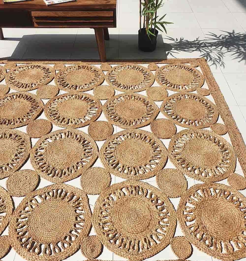 The RAMSHA HOME Braided Natural Outdoor Jute Rug (BR 086), featuring a woven sunburst pattern, adds eco-friendly luxury and durability to the tiled floor by a wooden table and potted plant as sunlight filters through nearby foliage.