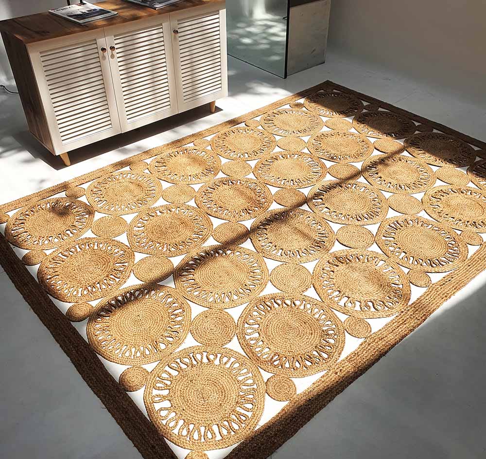 The RAMSHA HOME Braided Natural Outdoor Jute Rug (BR 086), featuring a sunburst design in brown and tan, sits on a light floor beside a white cabinet with wooden top, sunlight streaming into the minimalist room.