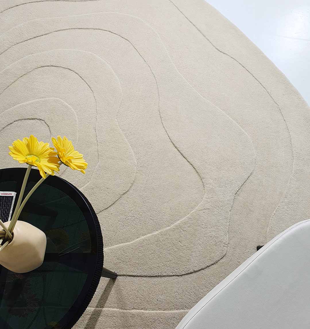 The RAMSHA HOME Luxurious Multi Color Round Handmade Rug - HTW 63 (200cm) features curved, raised lines like contour maps. On the rug sits a small black table with a vase of yellow flowers beside a white chair.