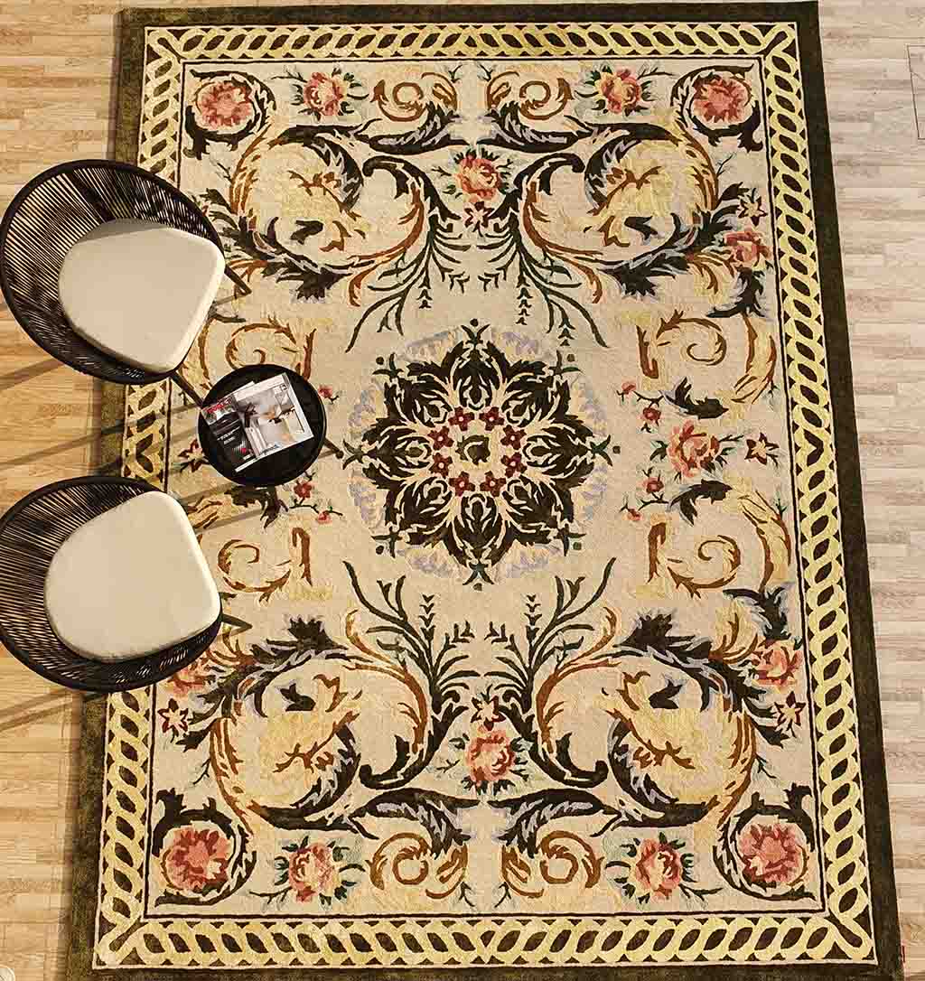 The Luxurious Wool, Silk & Cotton Handmade Rug - HTWV 03 (300x400cm) by RAMSHA HOME features intricate gold, green, and pink floral designs; two modern black chairs with white seats and a small round table rest on the rug.