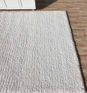 The RAMSHA HOME Soft White Rectangle Bedroom Shaggy Rug (SG 06) features a plush texture and is placed on a light brown, wood-patterned tile floor near a white cabinet, with sunlight highlighting its soft pile.