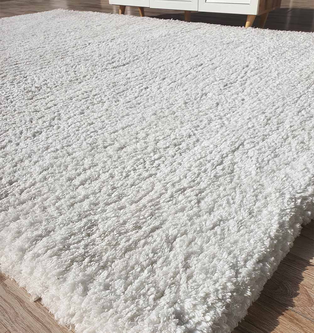 The RAMSHA HOME Soft White Rectangle Bedroom Rug - SG 06 is a shaggy rug with a pebbled texture and fluffy pile, shown close up on a wooden floor in a bright, modern room.