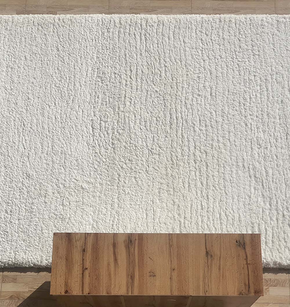 The RAMSHA HOME Soft White Rectangle Bedroom Rug Shaggy Rug - SG 06 with a pebbled shag texture lies on a wooden floor. A natural-finish wooden table is partially visible at the bottom of the image.