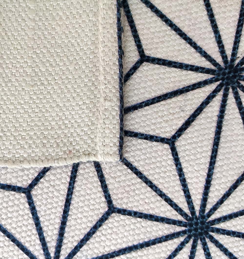 A close-up of two overlapping fabrics: one is plain light beige, while the other is RAMSHA HOME’s Geometry Pattern Printed Durry in white and black (140x200cm, DD-006) with a bold geometric starburst design.
