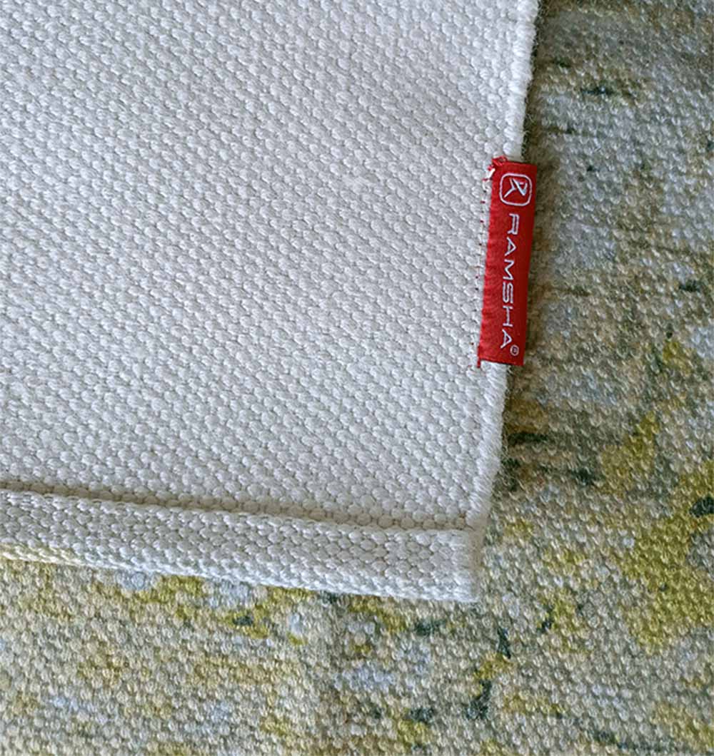 A close-up of the corner of the Geometry Pattern Printed Durry Green Color DD-009 by RAMSHA HOME, featuring a textured white modern abstract design and a red RAMSHA HOME label, on a green and yellow patterned surface.