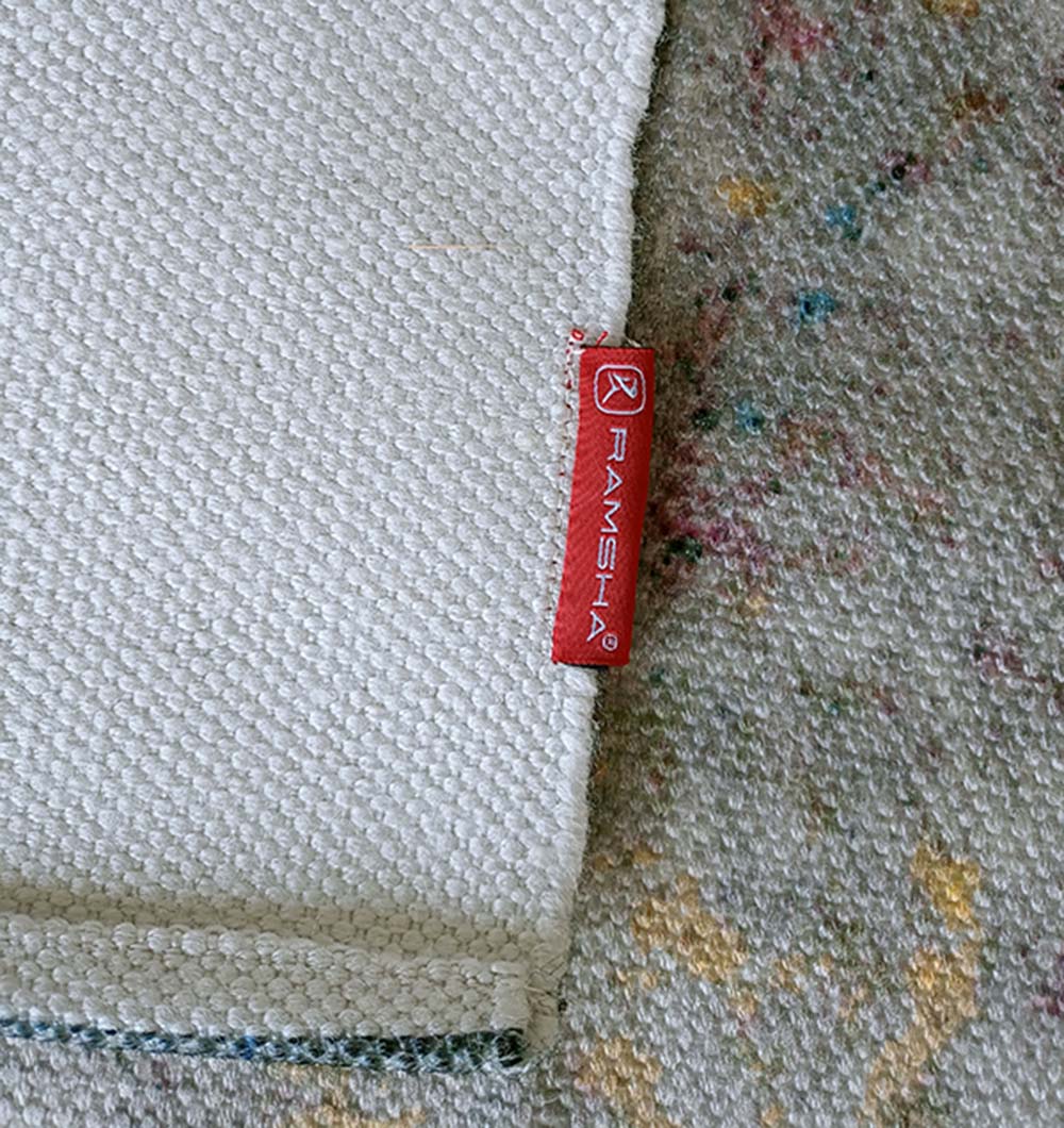 A close-up shows the corner of a woven white rug with a red RAMSHA HOME tag, placed on the Geometry Pattern Printed Durry Multi Color DD-010, featuring modern abstract design elements.
