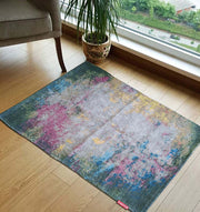 The Geometry Pattern Printed Durry Multi Color DD-010 by RAMSHA HOME adds vibrant style to the wooden floor near a large window, accented by a potted plant and brown armchair, while trees and a road outside enhance the rug’s modern look.