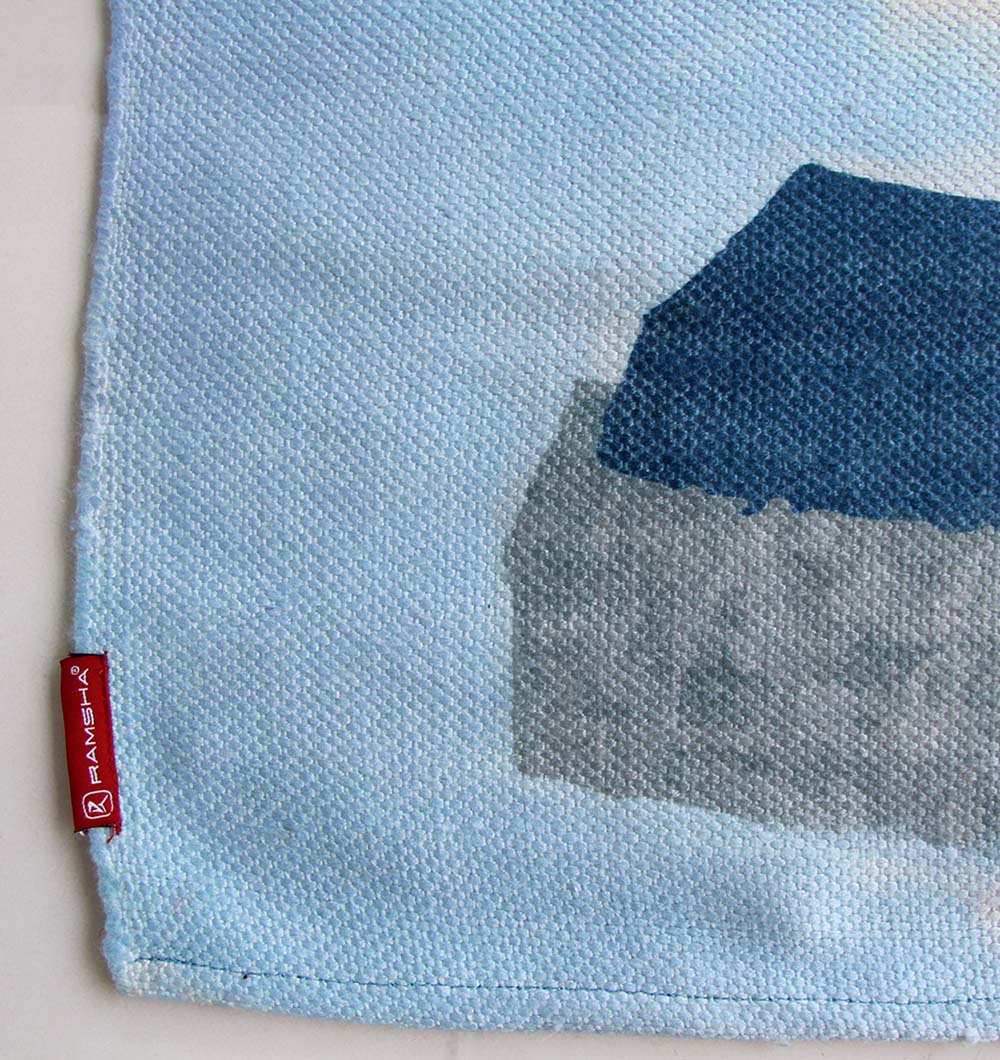A close-up of the Multi Color Decor Area Rug 150X210 cm DD-037 by RAMSHA HOME shows a blue and gray abstract pattern, with a small red tag featuring the RAMSHA HOME brand name sewn onto the edge.