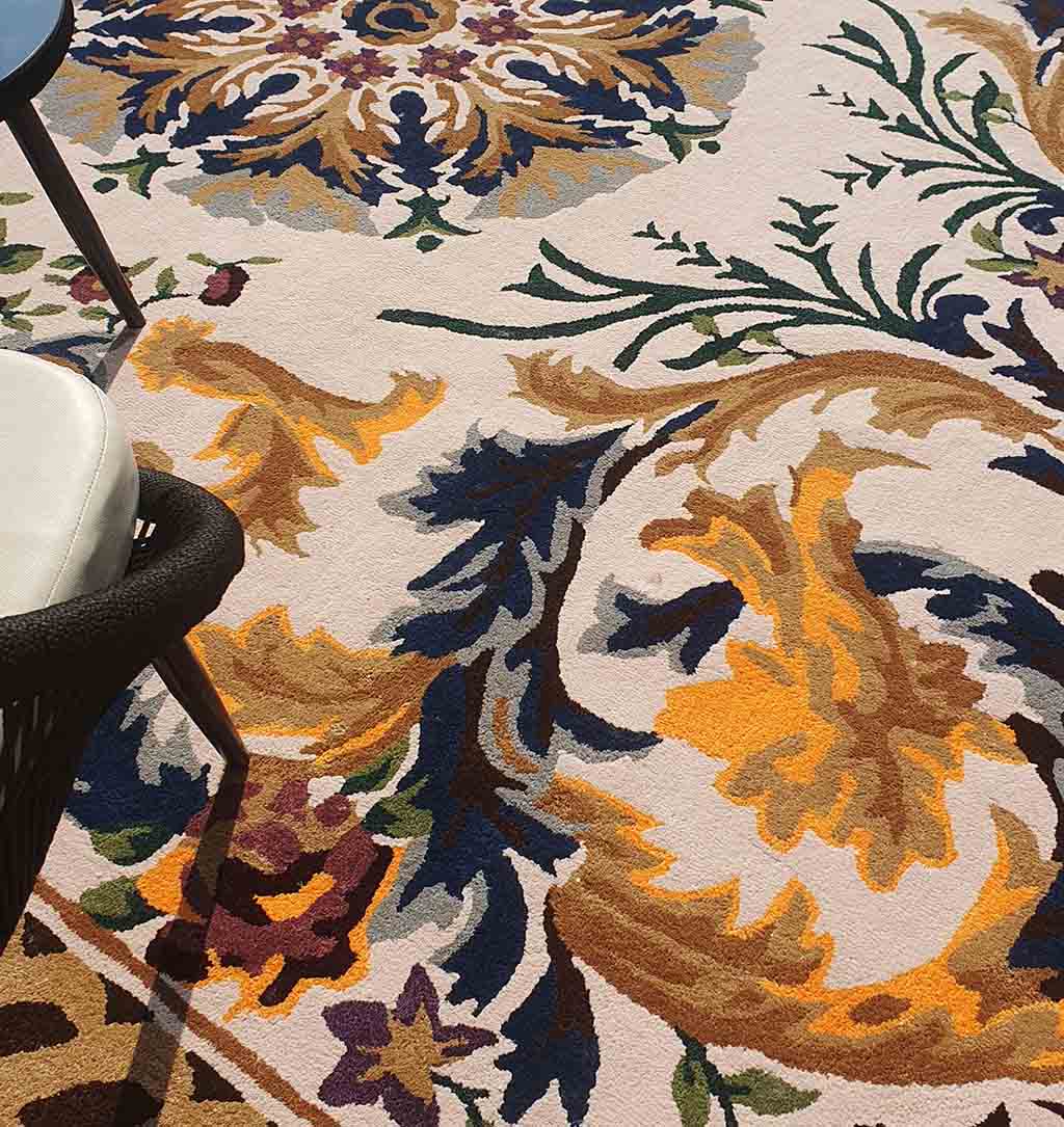 The RAMSHA HOME Polyester & Cotton Hall Decor Rug - HTP 02 (300x400cm) features ornate gold, blue, green, and purple floral patterns. Shown partially under a black chair with a white cushion, warm light highlights its vibrant design.