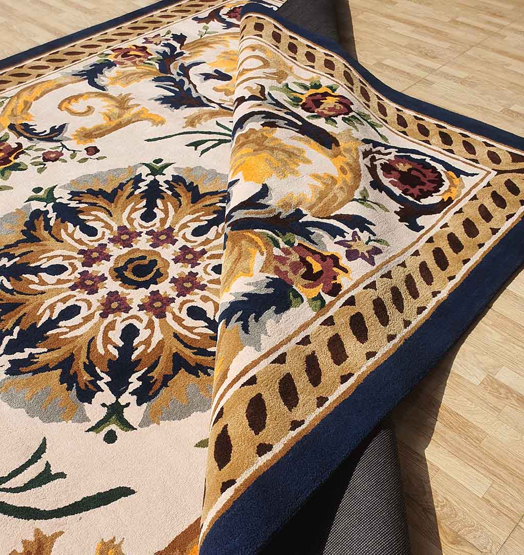 A close-up of the RAMSHA HOME Polyester & Cotton Hall Decor Rug - HTP 02 (300x400cm) showcases its ornate floral patterns in blue, green, and burgundy with a navy and gold border, partly folded on a light wood floor.