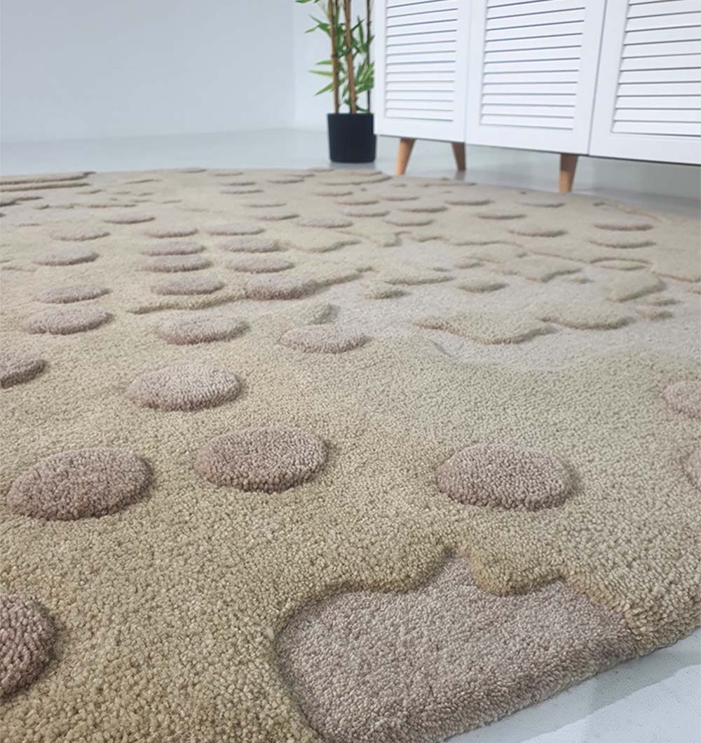 Close-up of the RAMSHA HOME Round Home Decor Handmade Carpet – HTW-56, featuring textured raised circular and abstract shapes, displayed on a light floor near a white cabinet and a potted plant in the background.